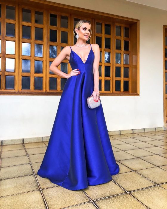 Mujer rubia con peinado de chongo usa vestido largo de fiesta color azul royal satinado con escote en v y tirantes, bolsa de mano brillante