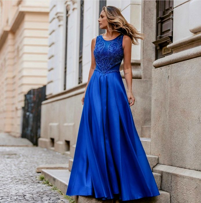 Mujer rubia parada en escalones con vestido de fiesta largo color azul royal sin mangas con ornamentos