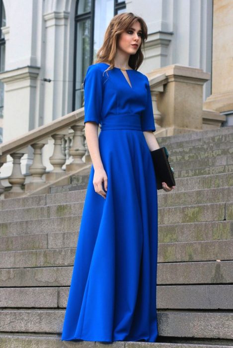 Chica de cabello castaño con fleco parada en escaleras usa vestido largo y sencillo color azul royal con mangas y cartera de mano