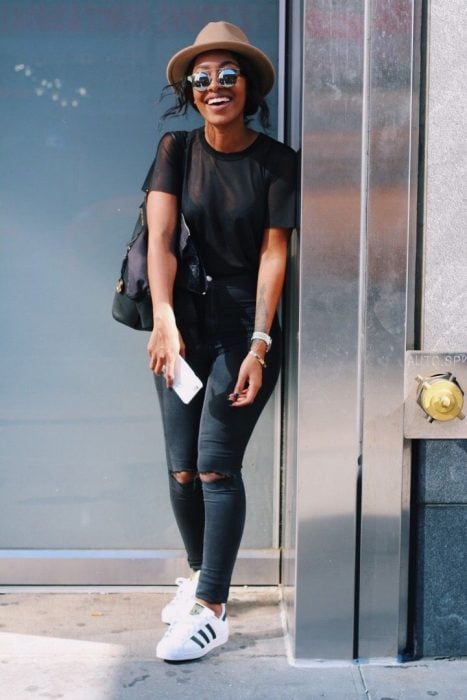  Chica recargada en pared usando pantalón negro rasgado, blusa negra, tenis blancos y sombrero café cálido 