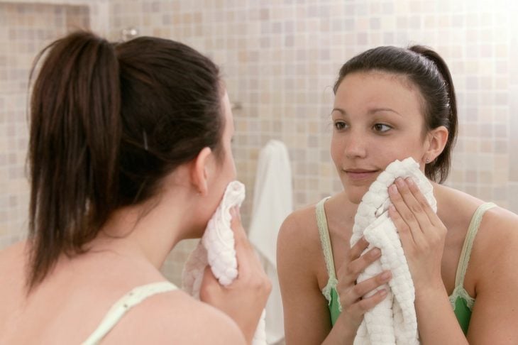Chica frente al espejo, usando una toalla blanca para secar su rostro después de haberlo lavado 