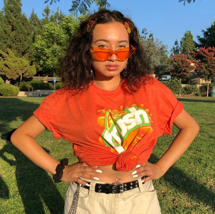 Chica con camiseta naranja, pantalón crema, lentes naranja transparente y broches en forma de flores