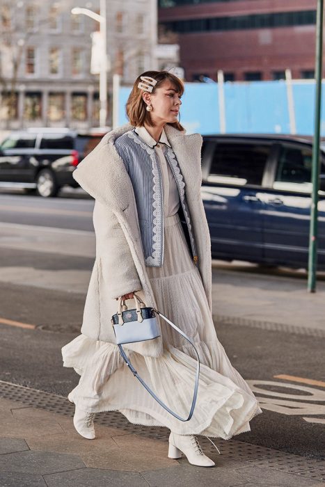 Chica caminando por la calle con vestido blanco de lana, saco, bolso en mano y un par de broches gigantes sobre su cabeza