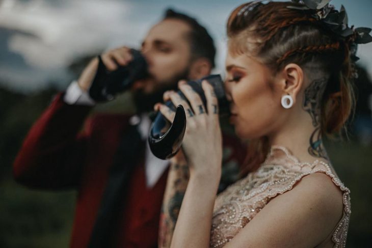 Pareja de novios celebra una boda nórdica al aire libre, novia con tatuajes en el cuello, brazos y manos, y expansiones y peinado de trenzas bebe hidromiel de cuerno vikingo