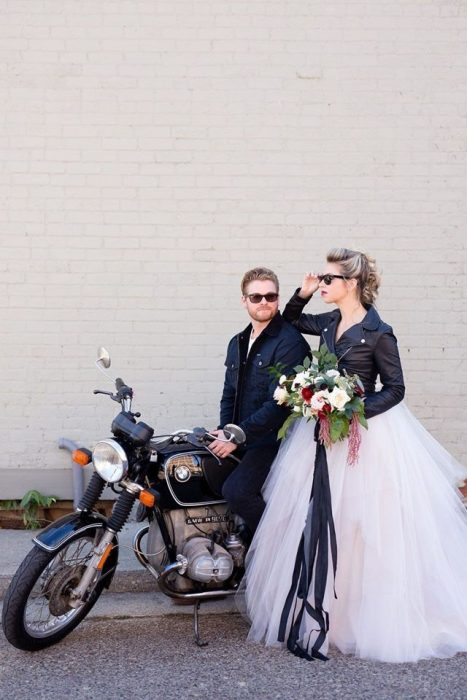 Novios recién casados usando chaquetas de cuero en color negro, recargados en una moto deportiva listos para llegar a su boda estilo biker 