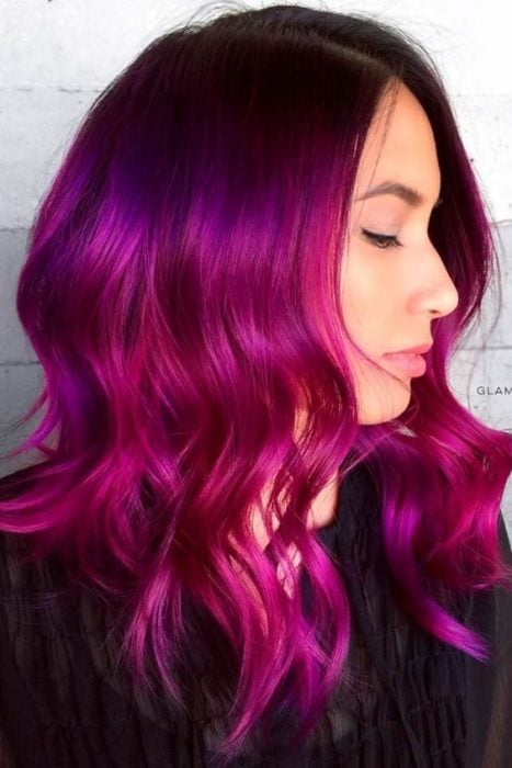 Chica recargada en una pared, volteada de perfil, llevando ropa oscura, mostrando su cabello bold pink con destellos purpuras 