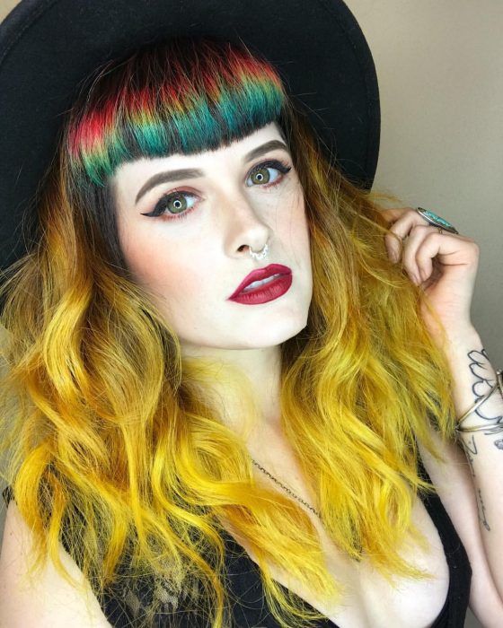 Chica con sombrero negro y cabello largo y ondulado color amarillo con fleco de colores rojo, amarillo y verde