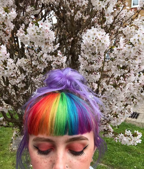 Chica con pecas y cavello lila con fleco de colores rojo, anaranjado, amarillo, verde, azul y morado frente a un árbol de flores blancas