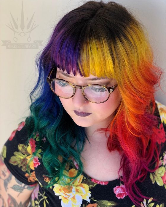 Chica de lentes retro con perforaciones en la nariz y vestido de flores, con cabello largo, ondulado y flequillo de colores del arcoíris, rosa, morado, azul, anaranjado, rojo, amarillo y verde