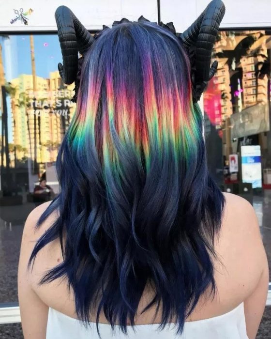 Chica con diadema de cuernos, cabello largo, ondulado, negro con una franda de colores arcoíris, morado, rojo, anaranjado, amarillo y verde 