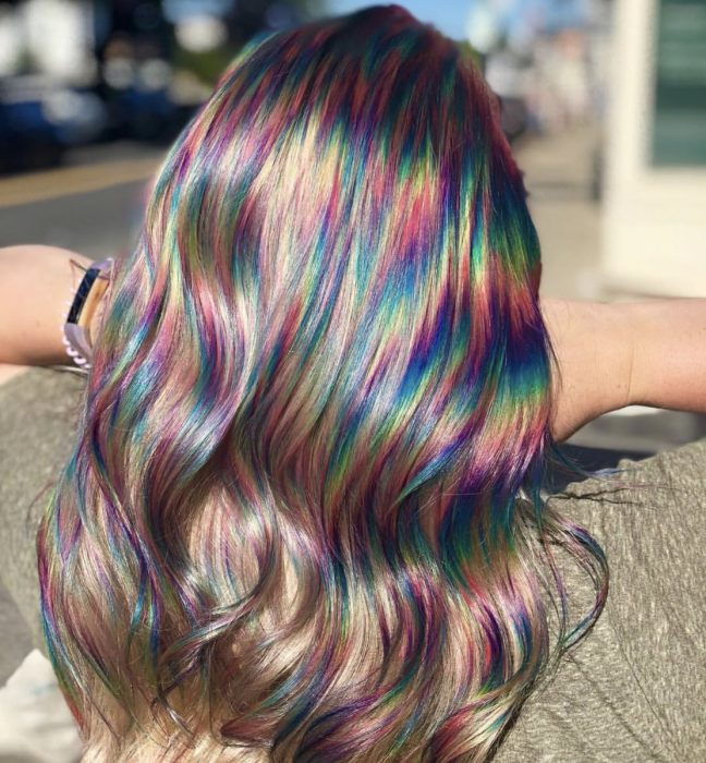 Mujer con cabello castaño rubio, ondulado con destellos de colores del aroíris, morado, rosa, amarillo, verde y azul