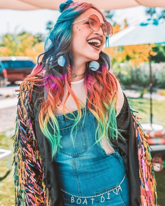 Chica con estilo hippie, overol de mezclilla, lentes rosas redondos, lentejuelas de estrellas en la cara simulando pecas, cabello largo ondulado de colores del arcoíris, azul, rosa, anaranjado, amarillo y verde, y aretes felpudos azules