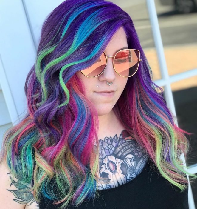 Chica con tatuaje en el pecho y perforaciones en la nariz y en el labio, con lentes rosas retro y cabello ondulado, largo y de colores del arcoíris, morado, azul, verde y rosa