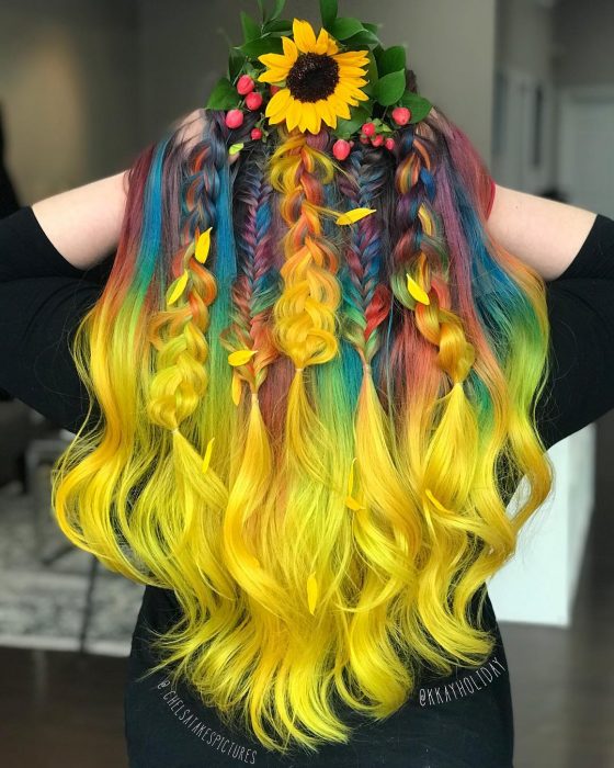 Chica con cabello largo y ondulado con trenzas, de colores del arcoíris, amarillo, verde, azul, anaranjado y morado, con una corona de girasol