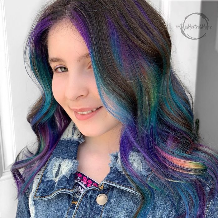 Tendencia de cabello, tinte oil slick que parece aceite derramado en el suelo, chica sonriendo con cabello largo, ondulado y de colores morado, azul, verde, rosa y amarillo