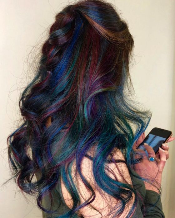 Tendencia de cabello, tinte oil slick que parece aceite derramado en el suelo, chica revisando su celular con cabello largo, ondulado peinado con una media trenza, de colores azul, morado, verde rosa y anaranjado