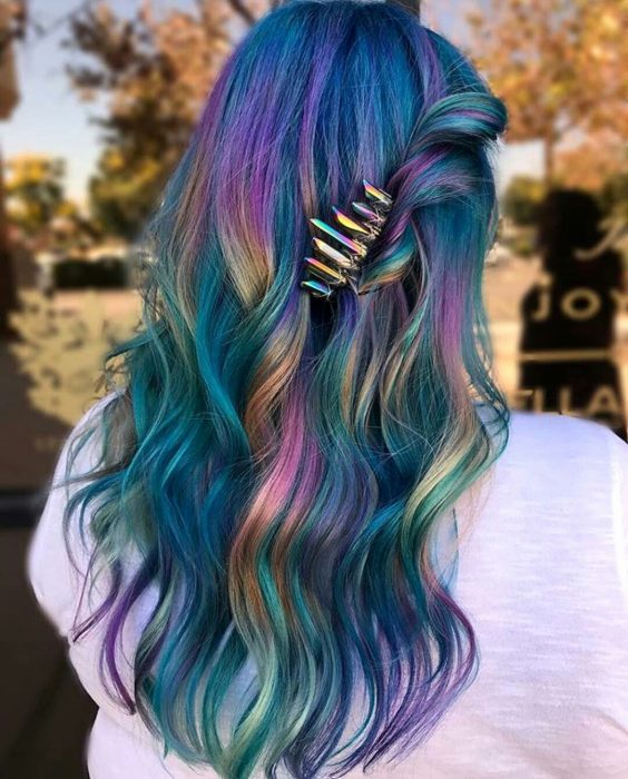 Tendencia de cabello, tinte oil slick que parece aceite derramado en el suelo, chica con cabello largo y ondulado de colores azul, morado, verde, rosa y anaranjado, con un broche tornasol