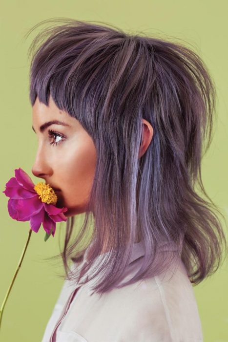 Chica de perfil oliendo una flor rosa con centro amarillo, cabello en capas hasta los hombros color gris lavanda o lavender gray