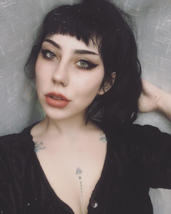 Chica parecida a Lady Gaga, de ojos verdes con delineado cat eye grueso y cabello negro oscuro tono inky black, perforación en la nariz y tatuajes en el pecho