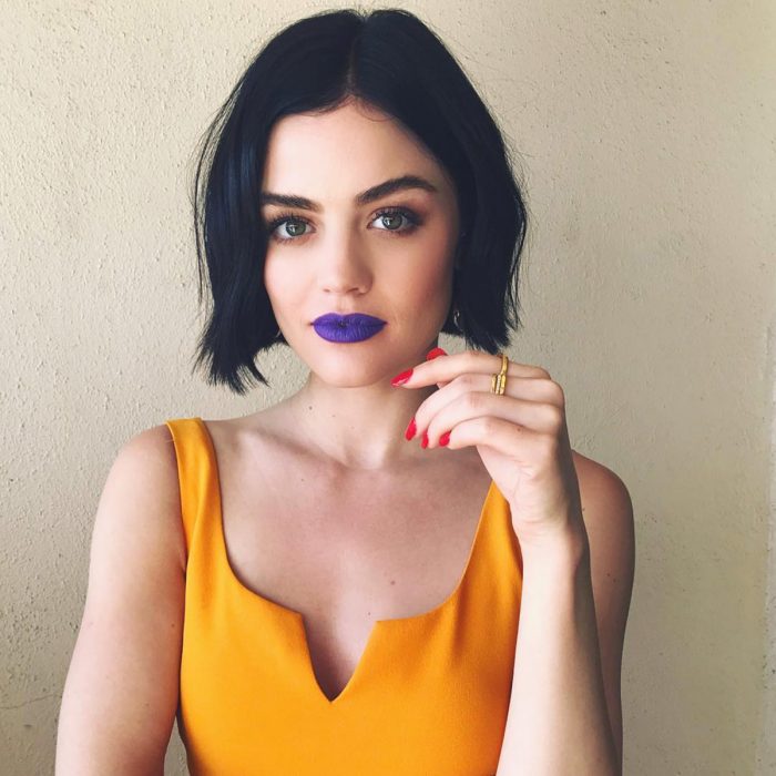 Actriz Lucy Hale, chica de ceja poblada, ojos verdes y labial morado, con cabello negro oscuro tono inky black corto arriba de los hombros, uñas rojas y vestido amarillo