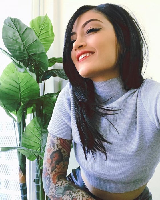 Matu Garcés. chica sonriendo frente a una maceta, tiene cabello negro oscuro tono inky black largo y lacio y tatuajes de arcoíris en los brazos