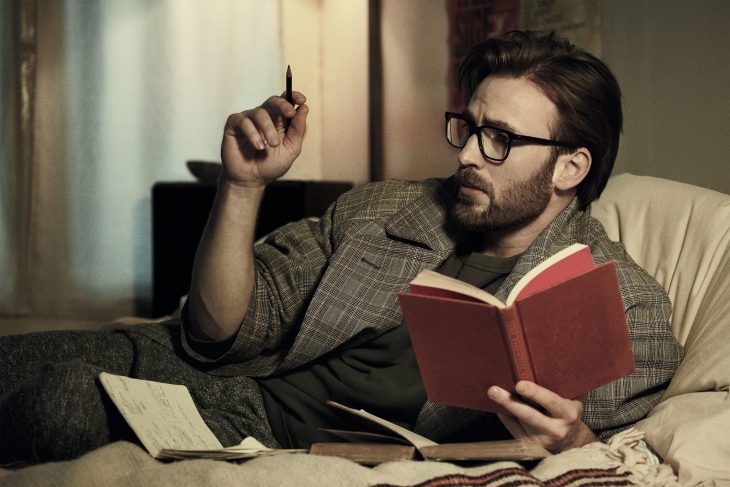 Chris Evans recostado en una cama con libros a su alrededor leyendo y escribiendo notas 