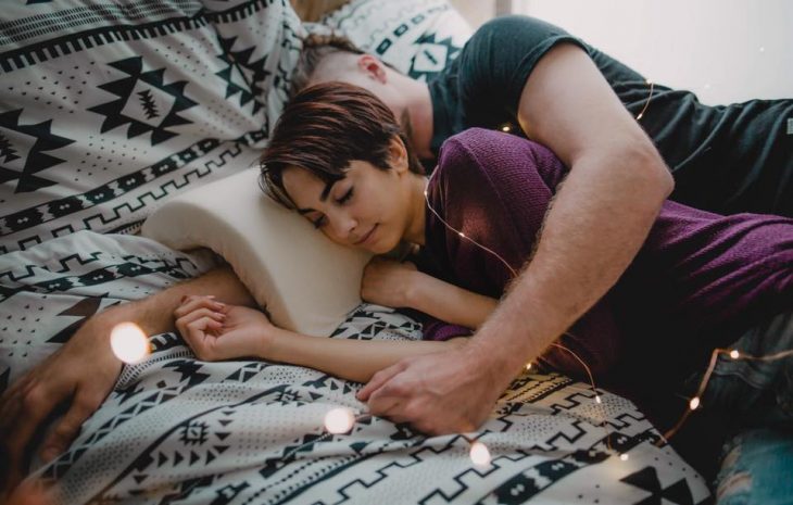 Pareja de novios recostados sobre una cama, abrazados de cucharita, usando la almohada Coodle Pillow