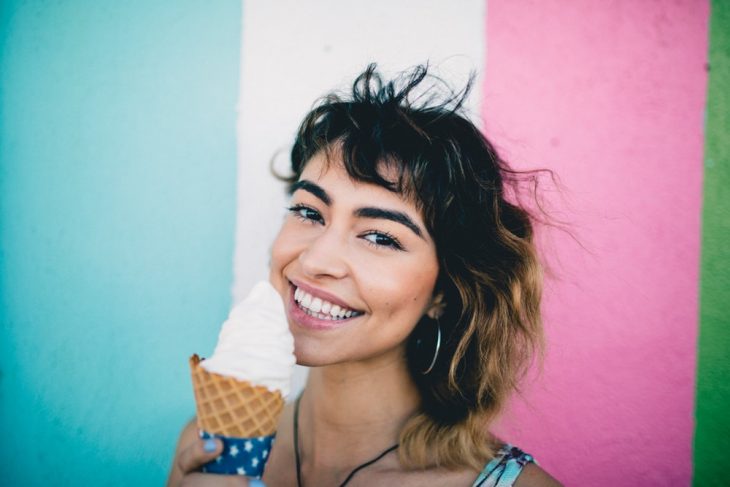 Chica comiendo helado de vanillina frente a una pared rayada a colores 