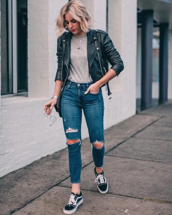 Chica caminando por la calle con jeans de mezclilla rotos, camisa gris, tenis negros con adornos en blancos y una chamarra de cuero