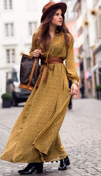 Chica caminando por la calle con un maxivestido color mostaza, botines negros, sombrero café y bolsa de mano