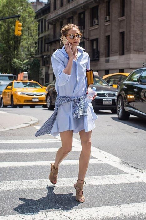 Chica caminando por la calle mientras habla por su celular, con vestido de camisa azul, camisa de cuadros amarrada a su cintura y gafas reflejantes