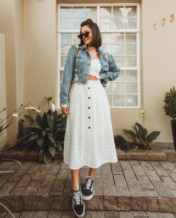 Chica modelando una falda larga blanca de lunares negros, un top blanco, chamarra de mezclilla y unos tenis negros con adornos en blanco y gafas oscuras