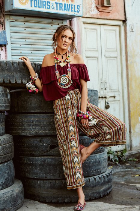 Chica en sesión fotográfica con pantalón con estampado holgado, blusa guinda holgada y gran variedad de accesorios estilo bohemio