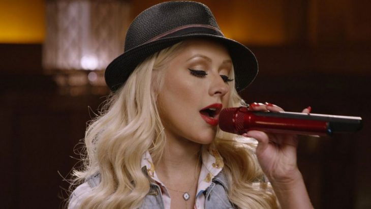 Christina Aguilera con sombrero gris, maquillaje dorado, sosteniendo un micrófono rojo, cantando