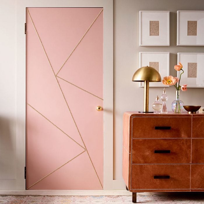 Puerta de una habitación que está junto a una vitrina y unos cuadros pintada de color rosa y decorada con líneas de color dorado 