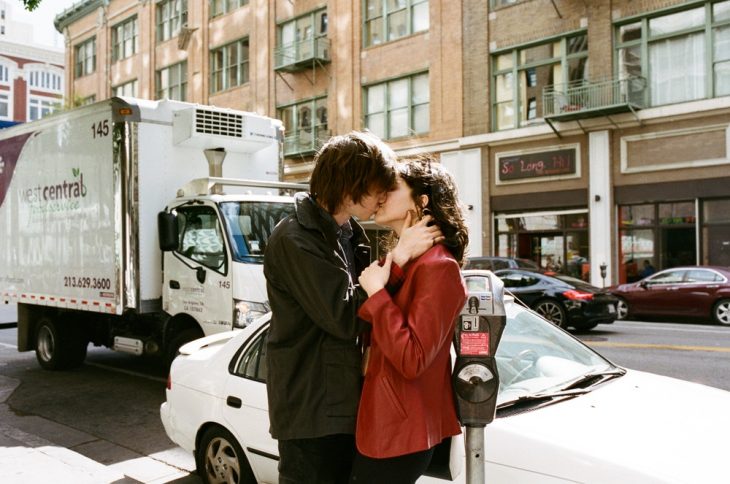Pareja de novios besándose apasionadamente a sobre la acera de una avenida transitada por autos