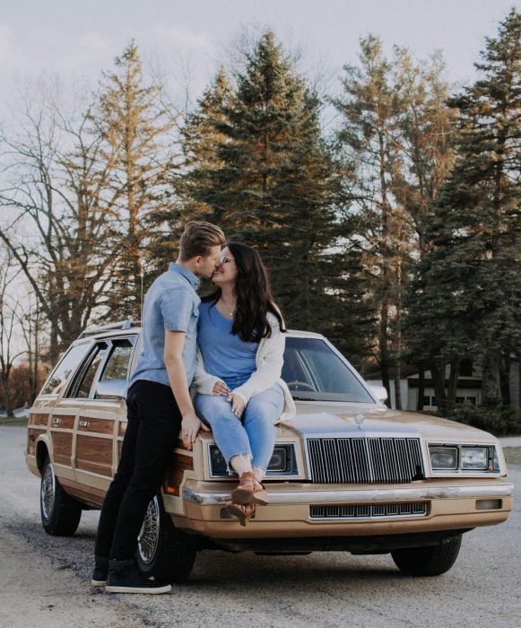 Chica sentada sobre el cofre de un automóvil, mientras es besada por su novio a mitad de carretera