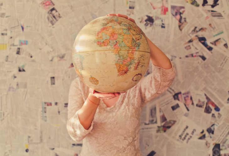 Mujer con blusa blanca de manga larga, sosteniendo un globo terraqueo entresus manos, cubriendo su rostro con el, mirando el país de escocia donde recetan viajes en la naturaleza 