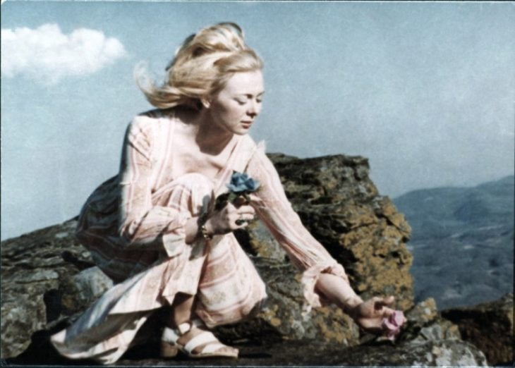 Joyce McKinney, chica parada sobre unas rocas a la orilla del mar usando maxivestido rosa con mangas largas, siendo golpeada por la brisa del mar, escena del documental Tabloid 