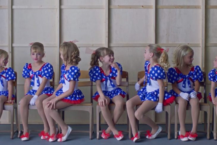  escena del documental Casting JonBenet, Grupo de niñas usando vestidos azul marino con puntos blancos, guantes blancos, zapatos rojos, sentadas en sillas, dentro de una sala de espera