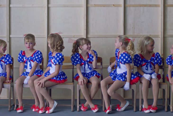  escena del documental Casting JonBenet, Grupo de niñas usando vestidos azul marino con puntos blancos, guantes blancos, zapatos rojos, sentadas en sillas, dentro de una sala de espera