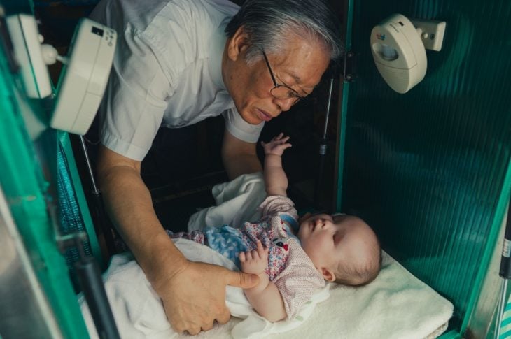 Hombre adulto con rasgos asiáticos inclinado para levantar a un bebé que se encuentra en un contenedor metálico, escena del documental The Drop Box 