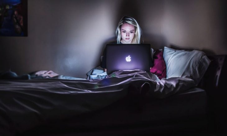 Chica sentada en una cama, con la laptop encima de sus piernas mirando series a media noche