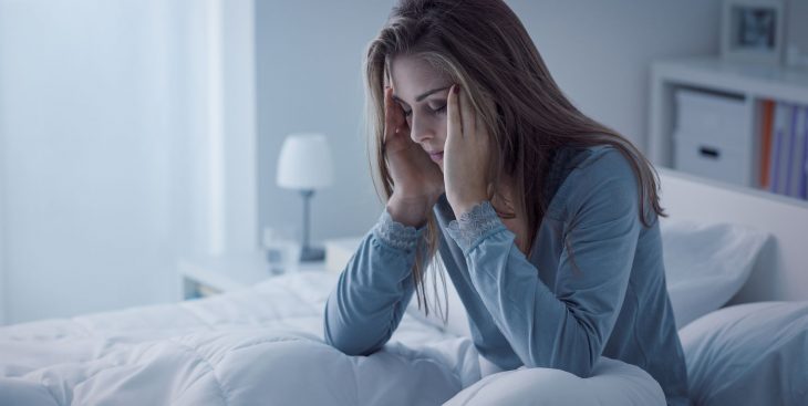 Chica tocando sus cienes con los ojos cerrados , la cara hacia abajo, con dolor de cabeza, sentada en una cama con sabanas bancas