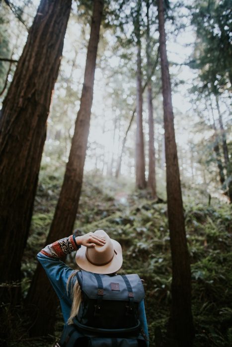 Chica parada en medio de un bosque sosteniendo su sombrero con una mano y admirando la naturaleza de escocia donde los doctores recetan viajes para sanar enfermos