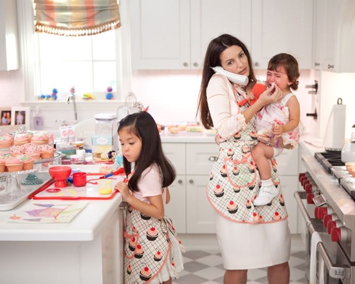 Madre sosteniendo a su bebé en brazos mientras habla por teléfono y su otra hija sobre la mesa viendo unos cupcakes 