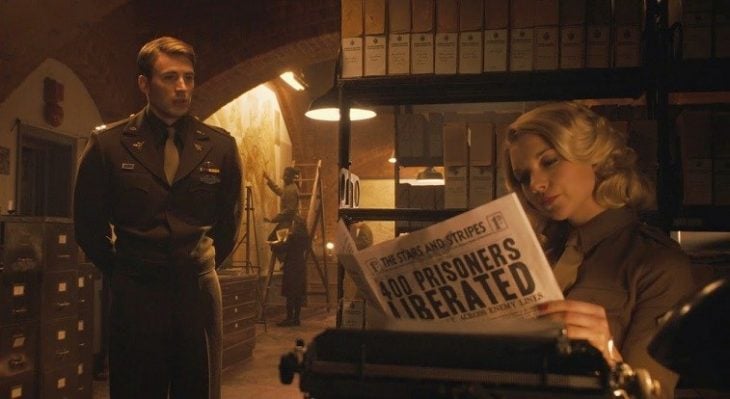 Chica sentada en un escritorio leyendo el periódico, mientras un hombre la observa fijamente, escena de la película Capitán América: el primer vengador, Natalie Dormer, Chris Evans 