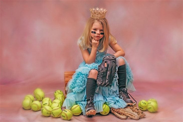 Niña con vestido azul estilo princesa sentada en una reja de madera rodeada de pelotas de tenis, fotografía por Heather Mitchell