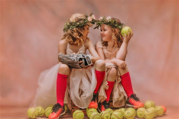 Niñas sentadas sobre una reja mirándose a los ojos, rodeadas de pelotas de tenis, usando vestido estilo princesa y calcetas deportivas, fotografía por Heather Mitchell