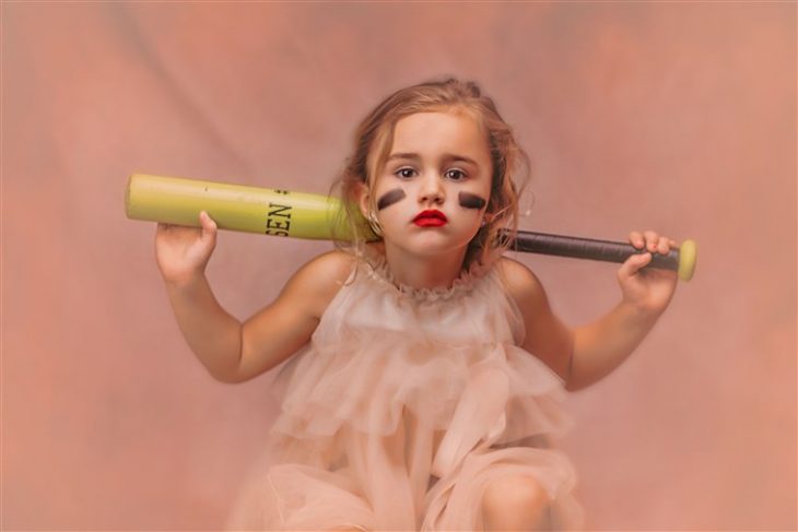 Niña sentada sobre un cajón de madera sosteniendo bat por la espalda, usando vestido estilo princesa fotografía por Heather Mitchell